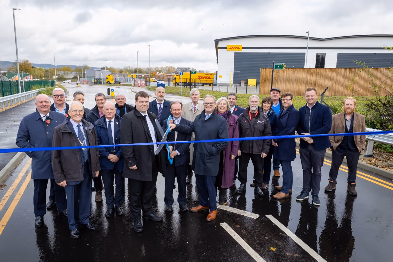Group of people in formal attire gathered outdoors for a ribbon-cutting ceremony near a DHL building.