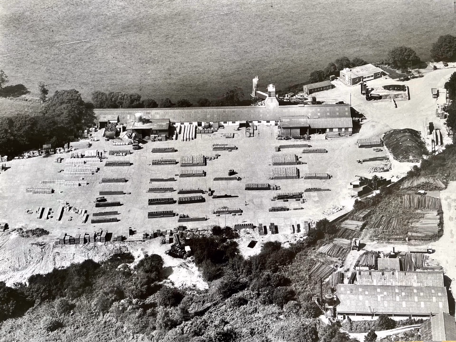 Aerial view of a rural area showing green fields, a parking lot with parked trucks, scattered buildings, and wooded areas extending to the horizon.