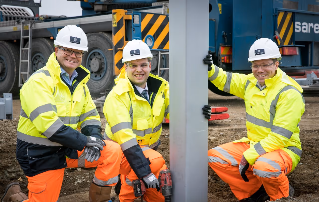 Three construction workers in high-visibility jackets and helmets smiling while kneeling around a steel beam on a construction site.