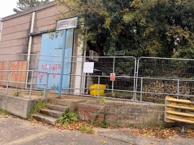 Entrance to Moulton Heights Goods with blue doors covered in red graffiti saying 'NO ENTR' and a drawing, behind metal railings and a temporary fence, with trees in the background.