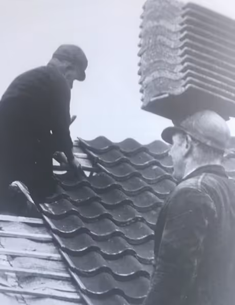 Two construction workers installing roof tiles on a sloped roof, one standing on the roof and the other looking on.