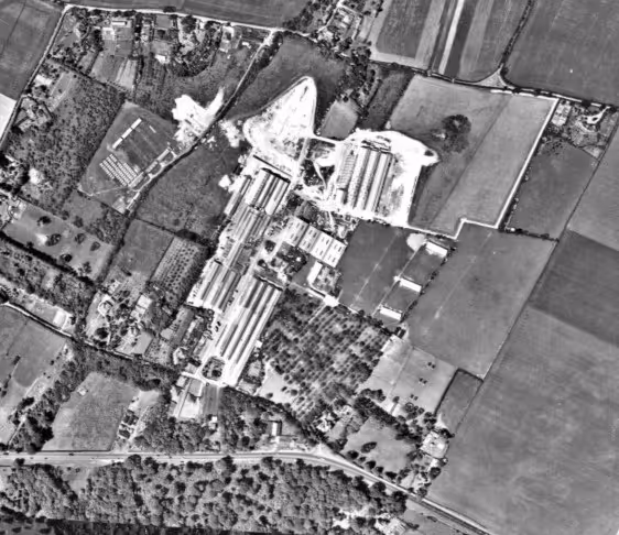 Black and white aerial photograph showing a rural area with agricultural fields, several large rectangular buildings, trees, and roads.