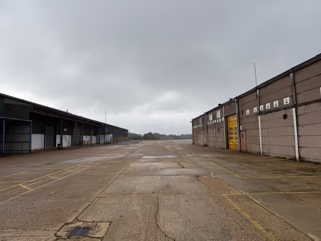 Empty industrial concrete yard flanked by warehouse buildings under a cloudy sky.