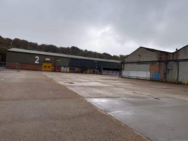 Empty industrial yard with two large warehouse buildings under cloudy sky.