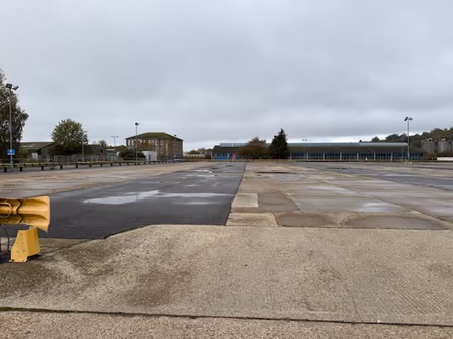 Empty concrete parking lot under a cloudy sky with buildings and trees in the background.
