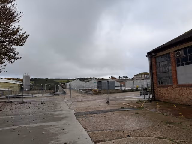 Wet industrial area with fenced-off greenhouses under a cloudy sky.
