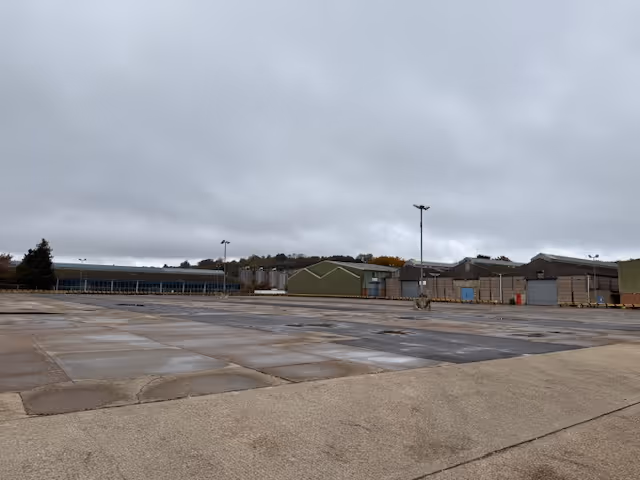 Empty industrial yard with concrete ground, several green and beige warehouse buildings under a cloudy sky.