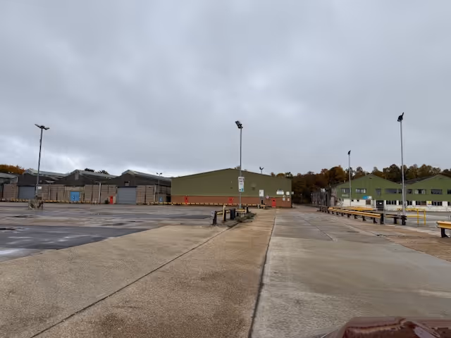 Empty industrial area with large green warehouses, concrete ground, and cloudy sky.