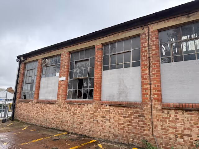 Brick building exterior with large windows, some broken and covered with boards, under an overcast sky.