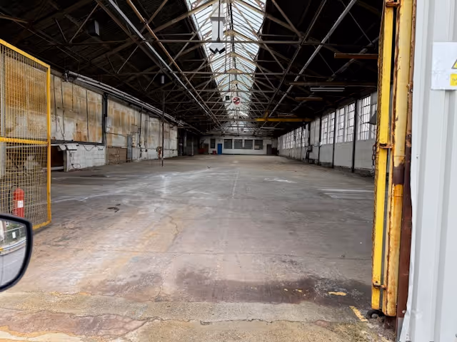 Empty industrial warehouse with large windows on the right and exposed roof trusses.