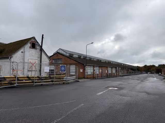 Long industrial warehouse building beside an empty wet asphalt road under a cloudy sky.