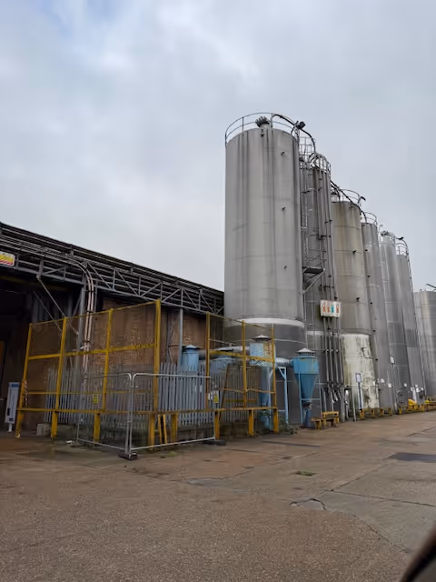 Tall industrial storage silos and metal structures outside a factory under a cloudy sky.