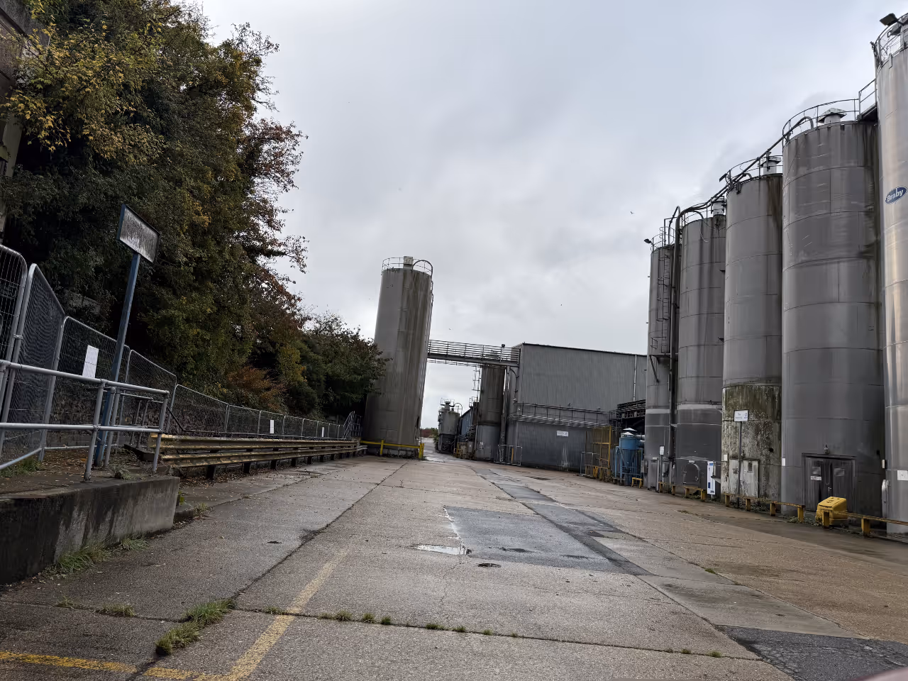 Industrial area with large metal silos on the right, a grey building ahead, and trees on the left under a cloudy sky.