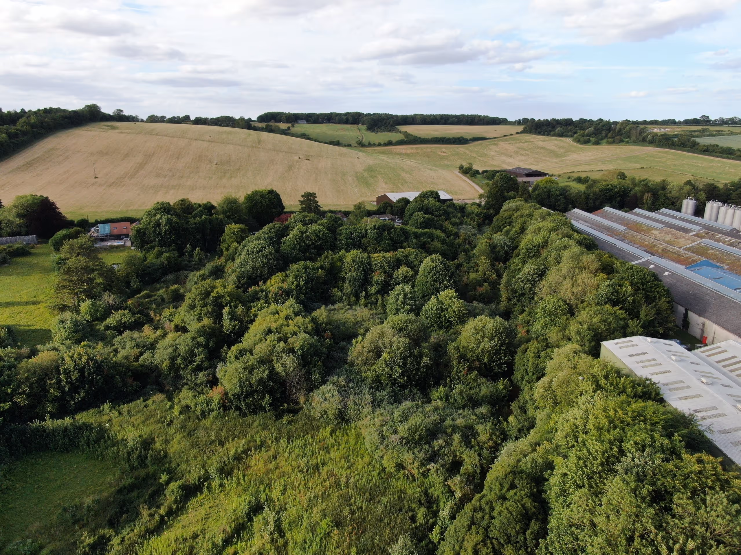 Aerial view of a lush green forest area adjacent to farmland and industrial buildings under a partly cloudy sky.