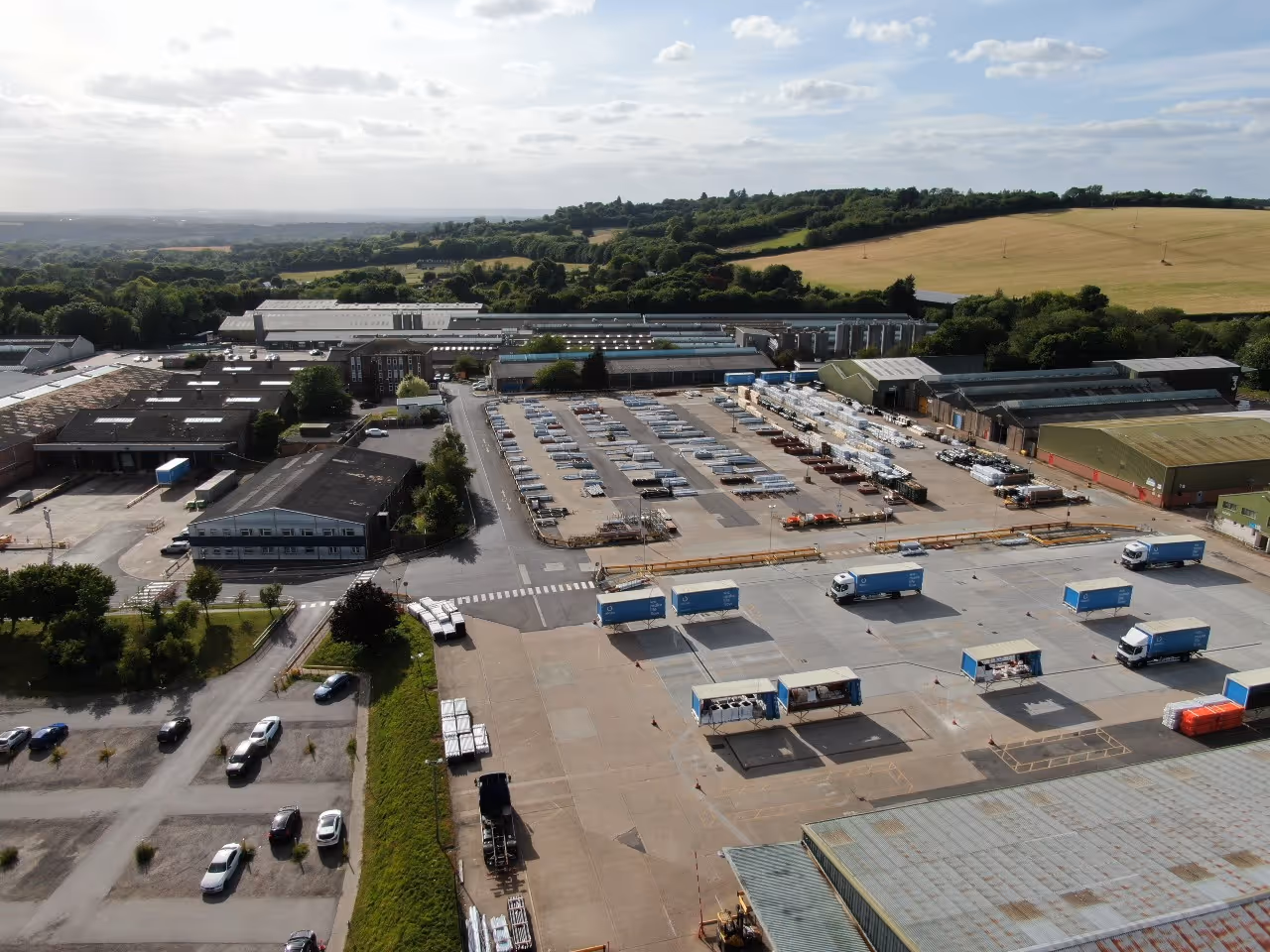 Aerial view of an industrial complex with multiple warehouses, parked trucks, and storage areas surrounded by green fields under a partly cloudy sky.