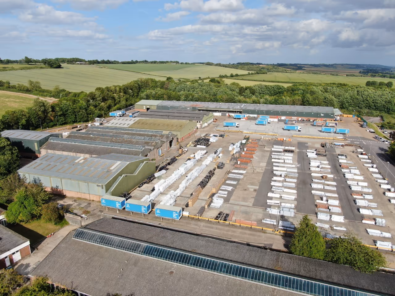 Aerial view of an industrial warehouse complex with multiple buildings, loading docks, parked blue trucks, and organized stacks of materials on a concrete yard surrounded by green fields and trees.