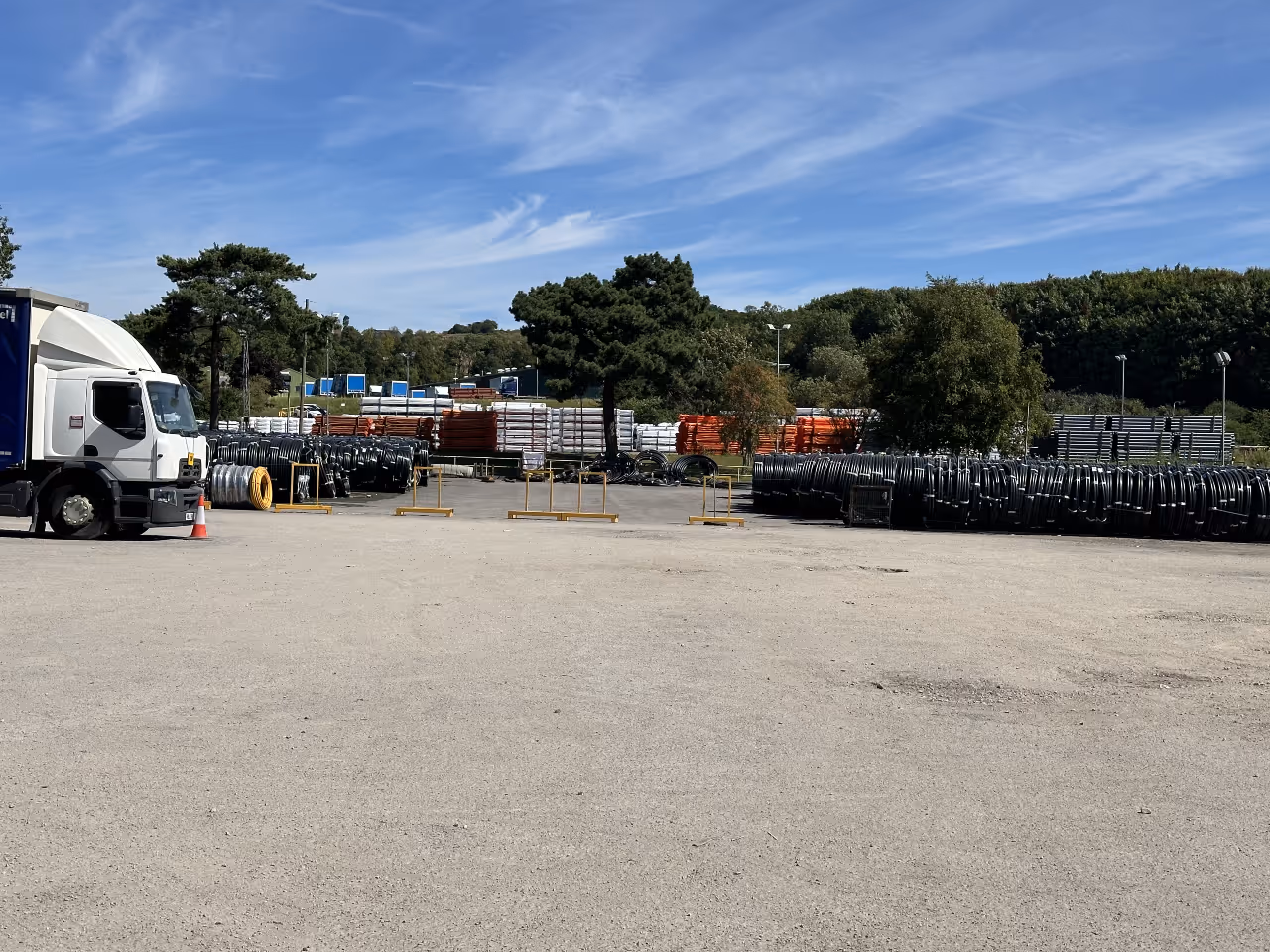 Industrial yard with stacks of coiled pipes, a parked white truck, trees, and blue sky with wispy clouds.