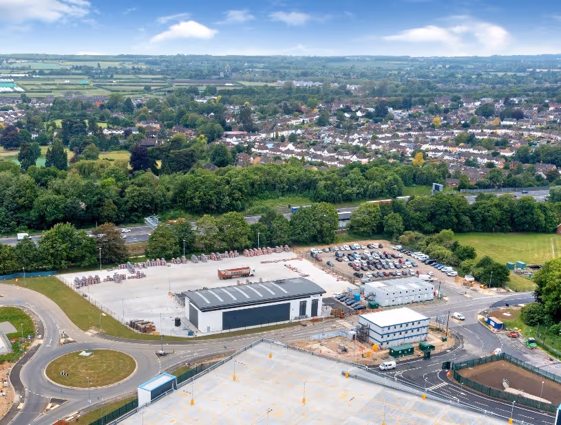 Aerial view of an industrial yard with stacked materials, several buildings, machinery, and surrounding greenery.
