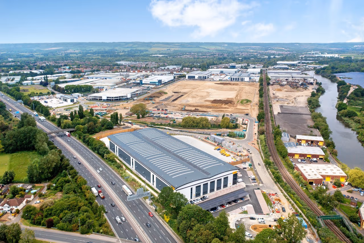 Aerial view of a highway with multiple lanes running beside industrial buildings, a large warehouse, railway tracks, and a river, under a partly cloudy sky.