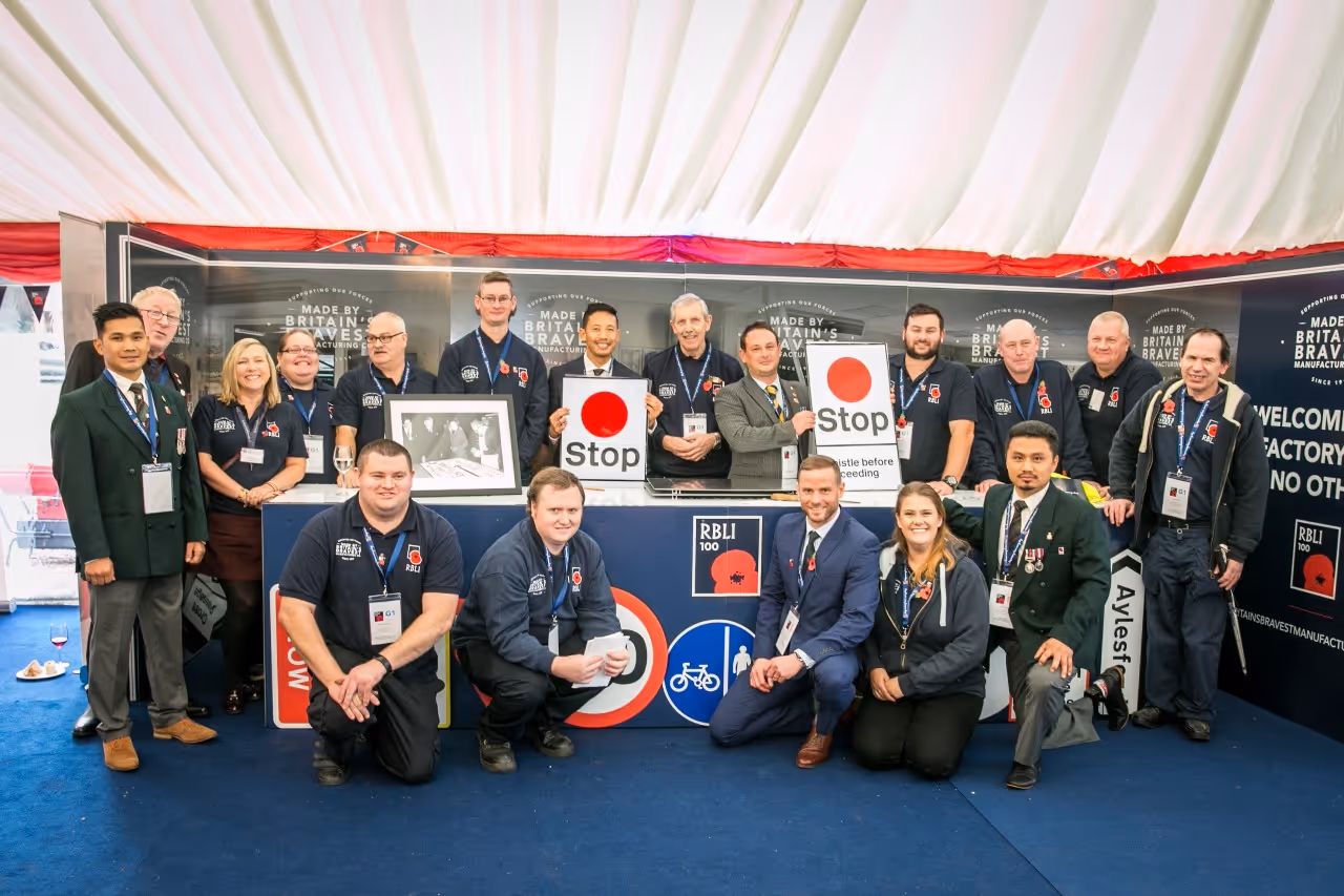 Group of people posing inside a tented exhibit with 'Made by Britain's Bravest' signage, some holding traffic stop signs and wearing poppy pins.
