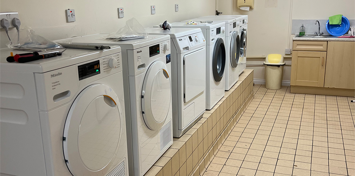 Row of white front-loading washing machines and dryers on a tiled platform in a laundry room.