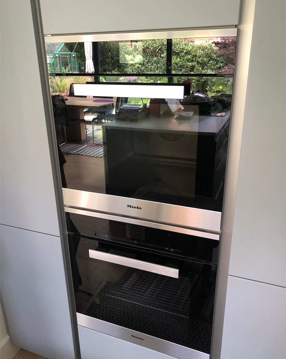 Modern kitchen with built-in stainless steel Miele double oven reflecting dining area and garden outside.