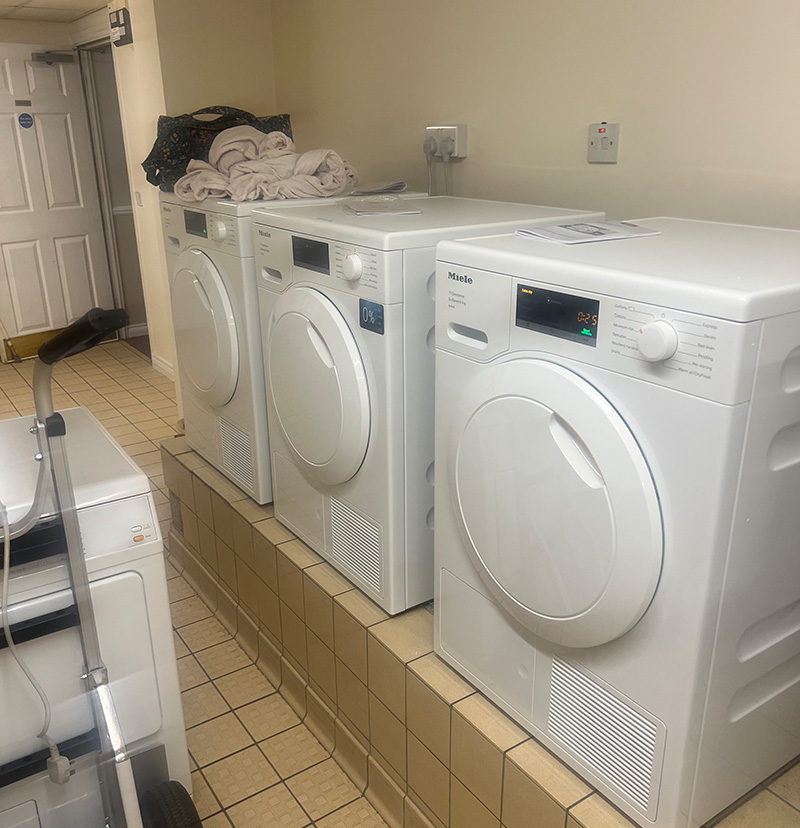 Three white Miele dryers lined up on a tiled platform in a laundry room with towels on top.