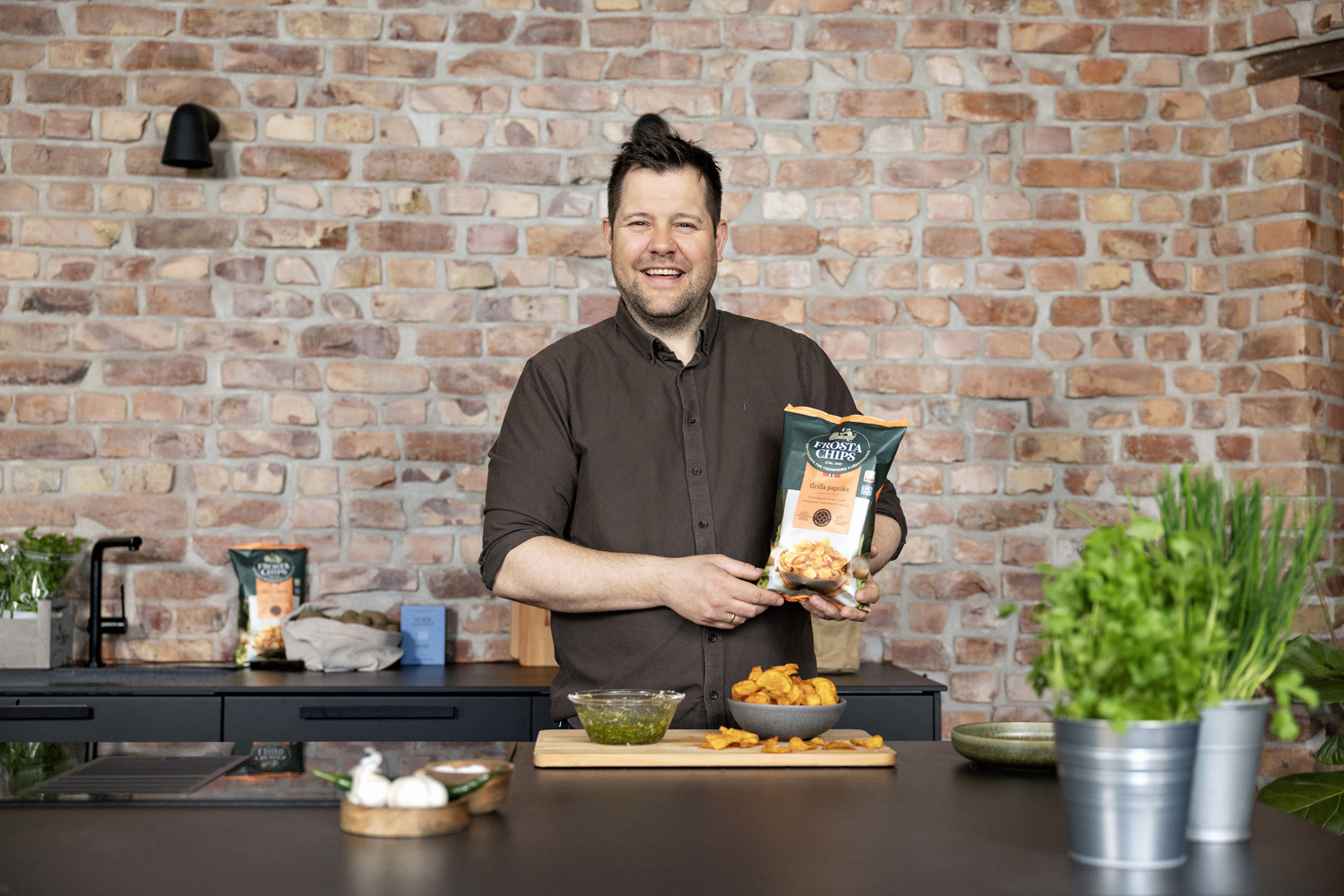 En kokk holder opp en pose chips i et rustik kjøkken, ser og smiler i kamera