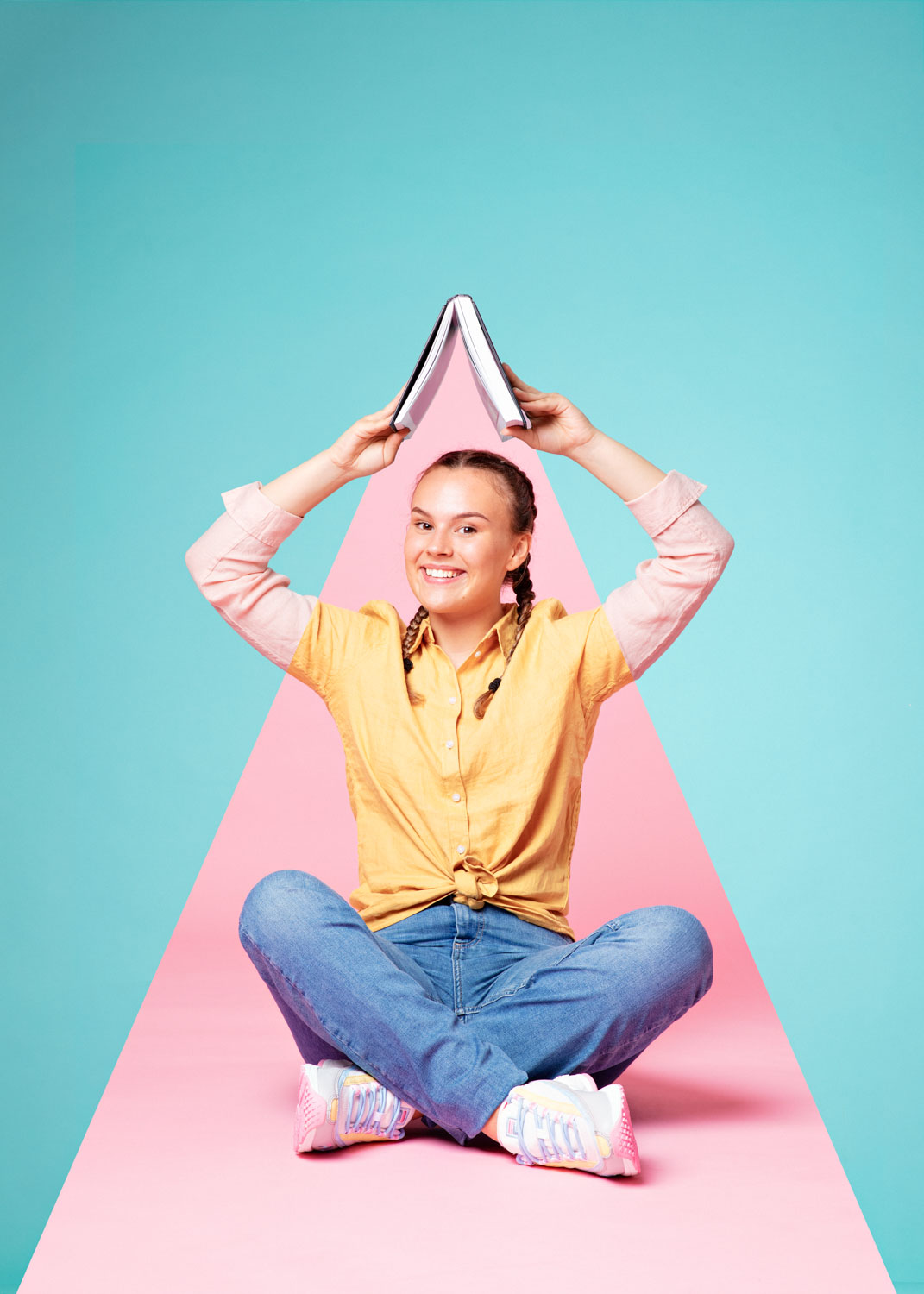 En ung kvinne sitter med beina i kryss mens huns holder en bok over hodet i et fargerikt fotostudio