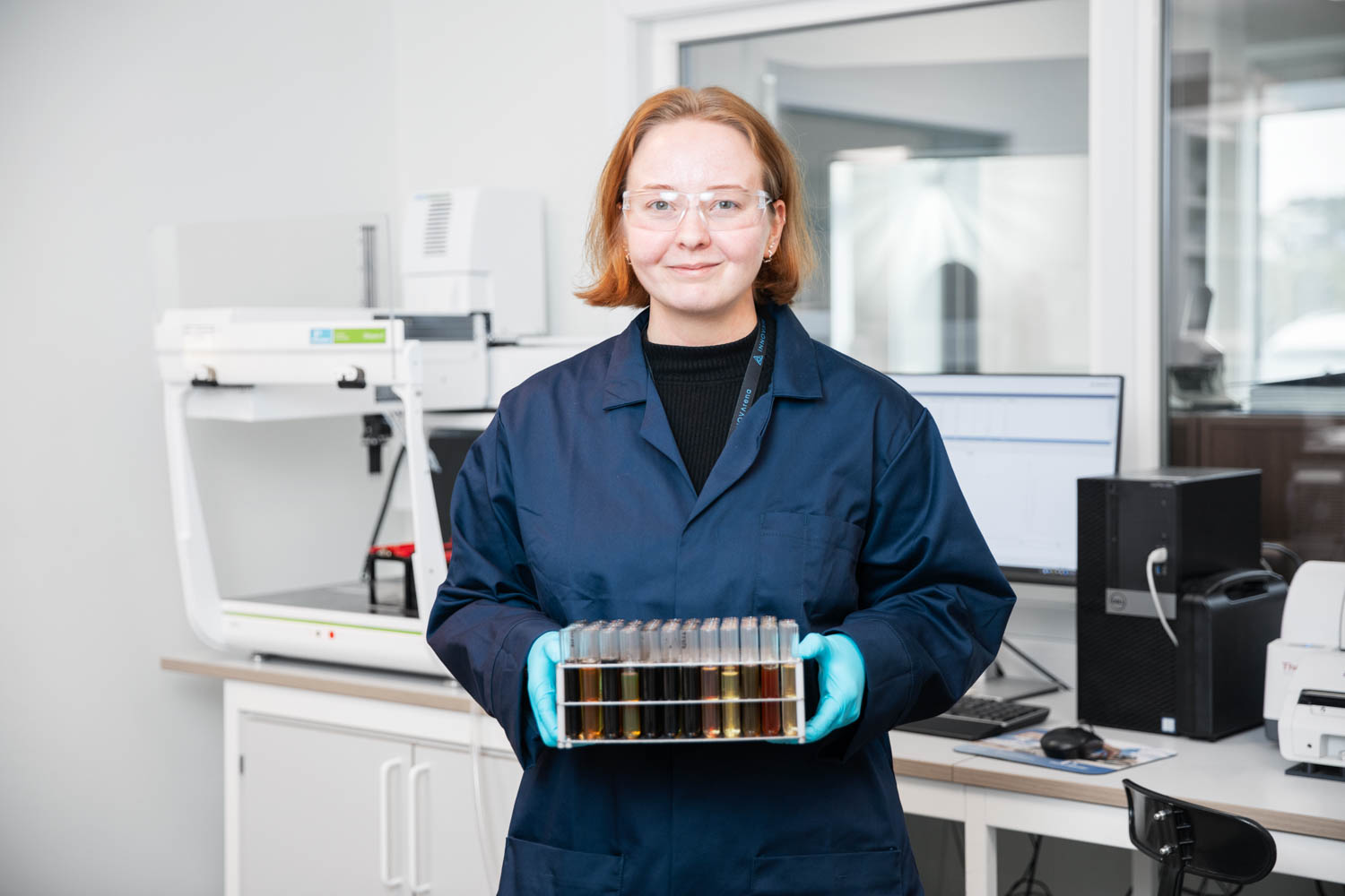 En ung kvinne i blå labuniform holder opp en rekke væskeprøver i en lab