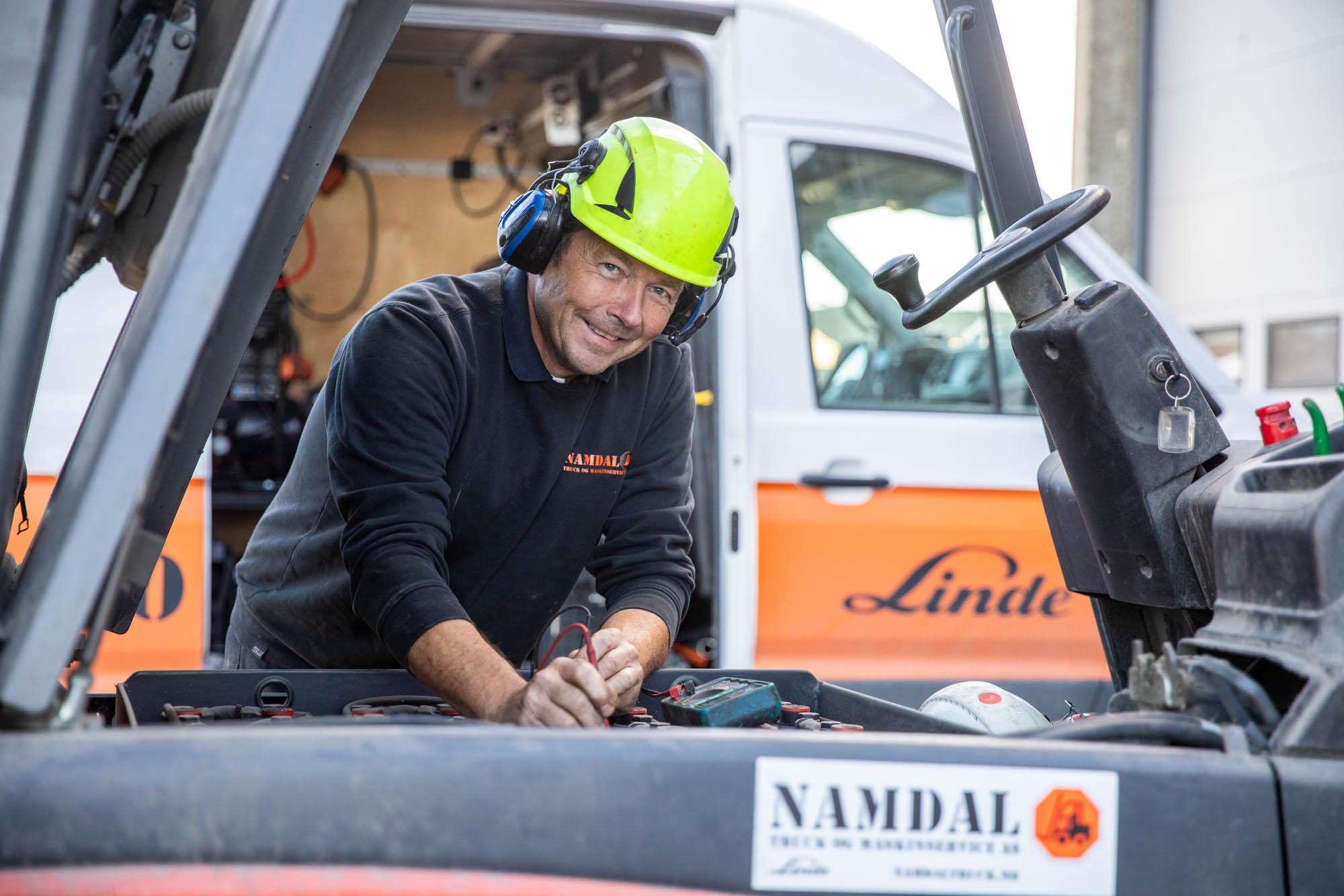 En mann i verneutstyr sitter inne i en truck og smiler til kamera