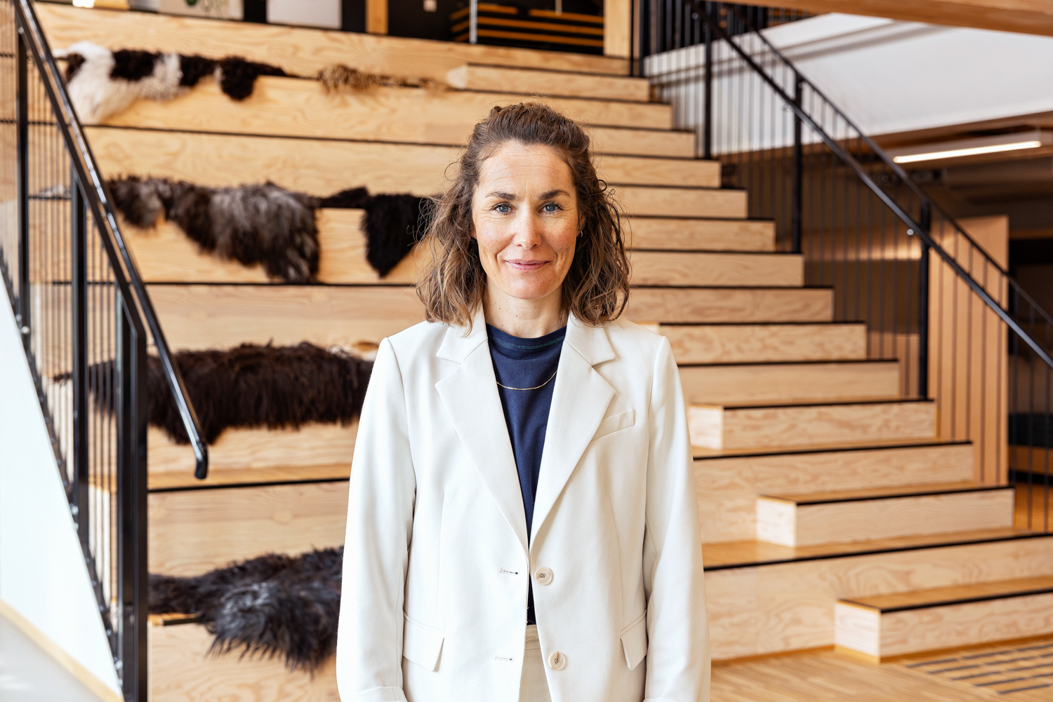 En kvinne i hvit dress ser rett i kamera mens hun står foran en tribune-trapp i et moderne lokale