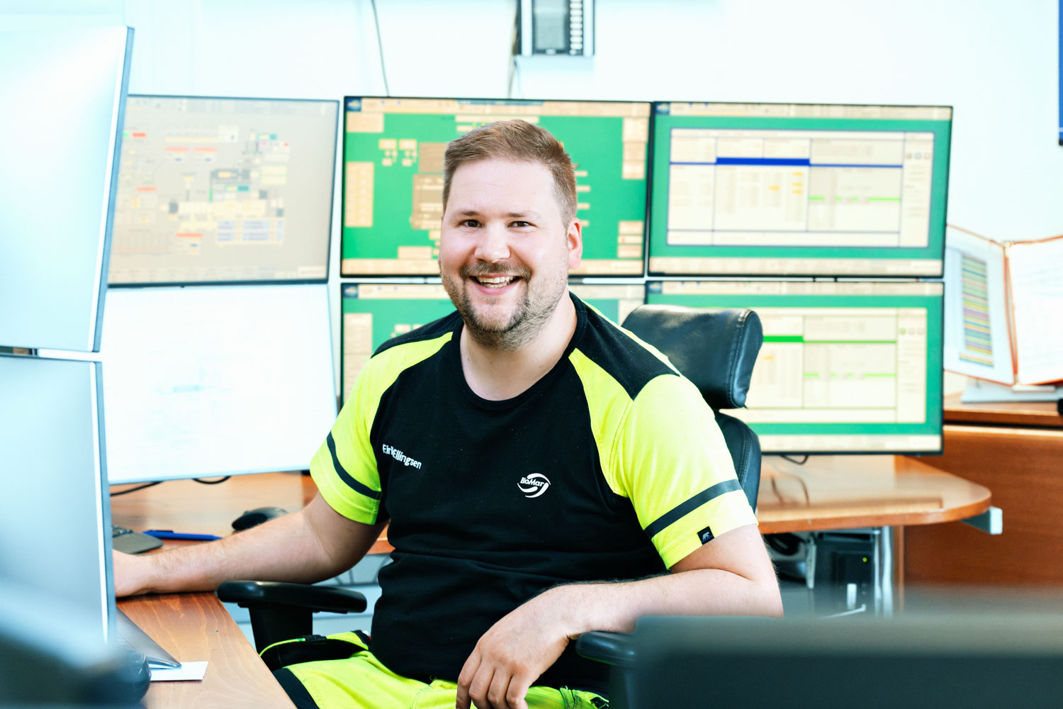 En mann iført synlighetsklær sitter inne på et kontor med flere monitorer på pulten