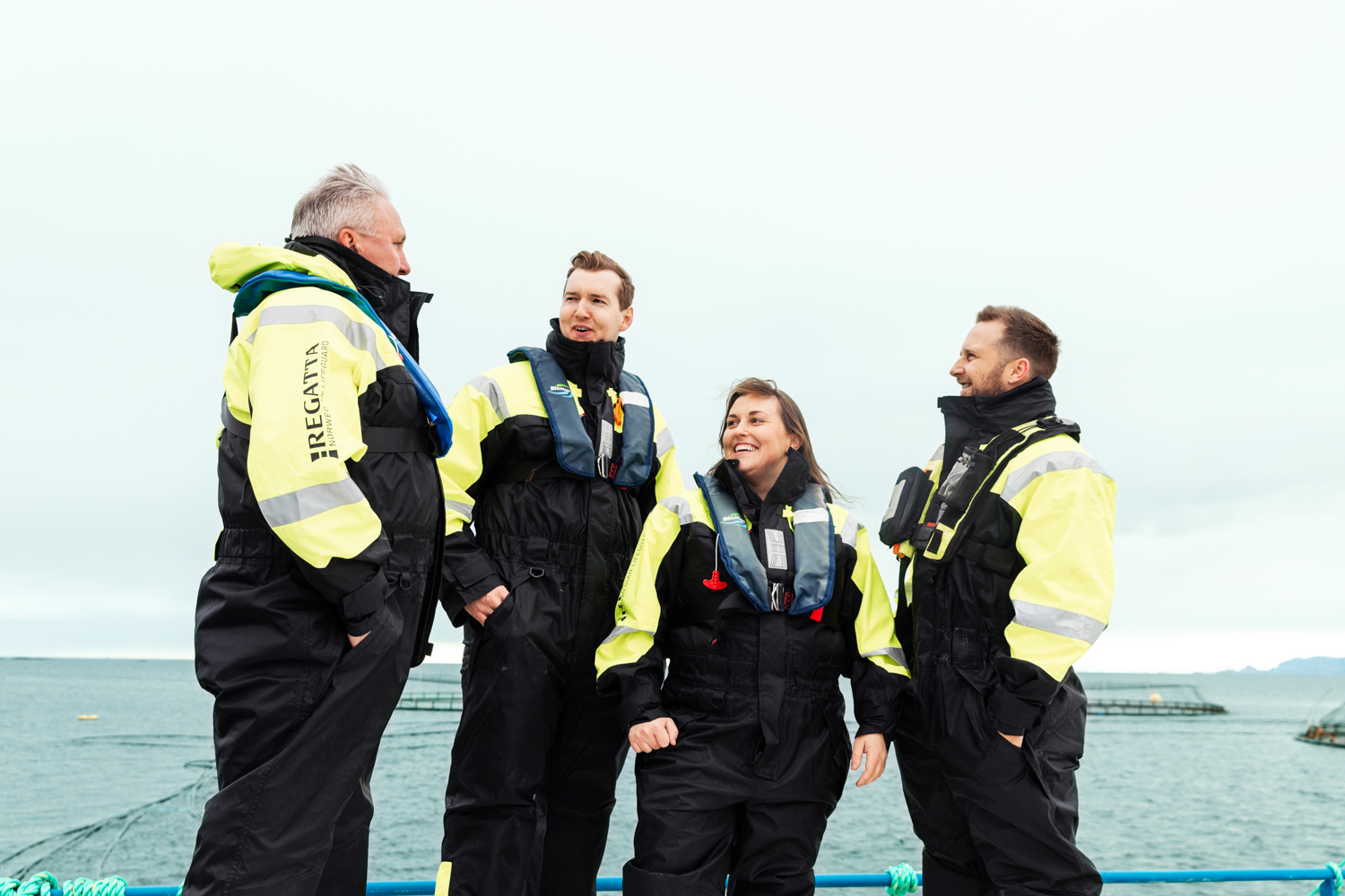 Fire personer iført synlighetsklær står og smiler til hverandre foran havet