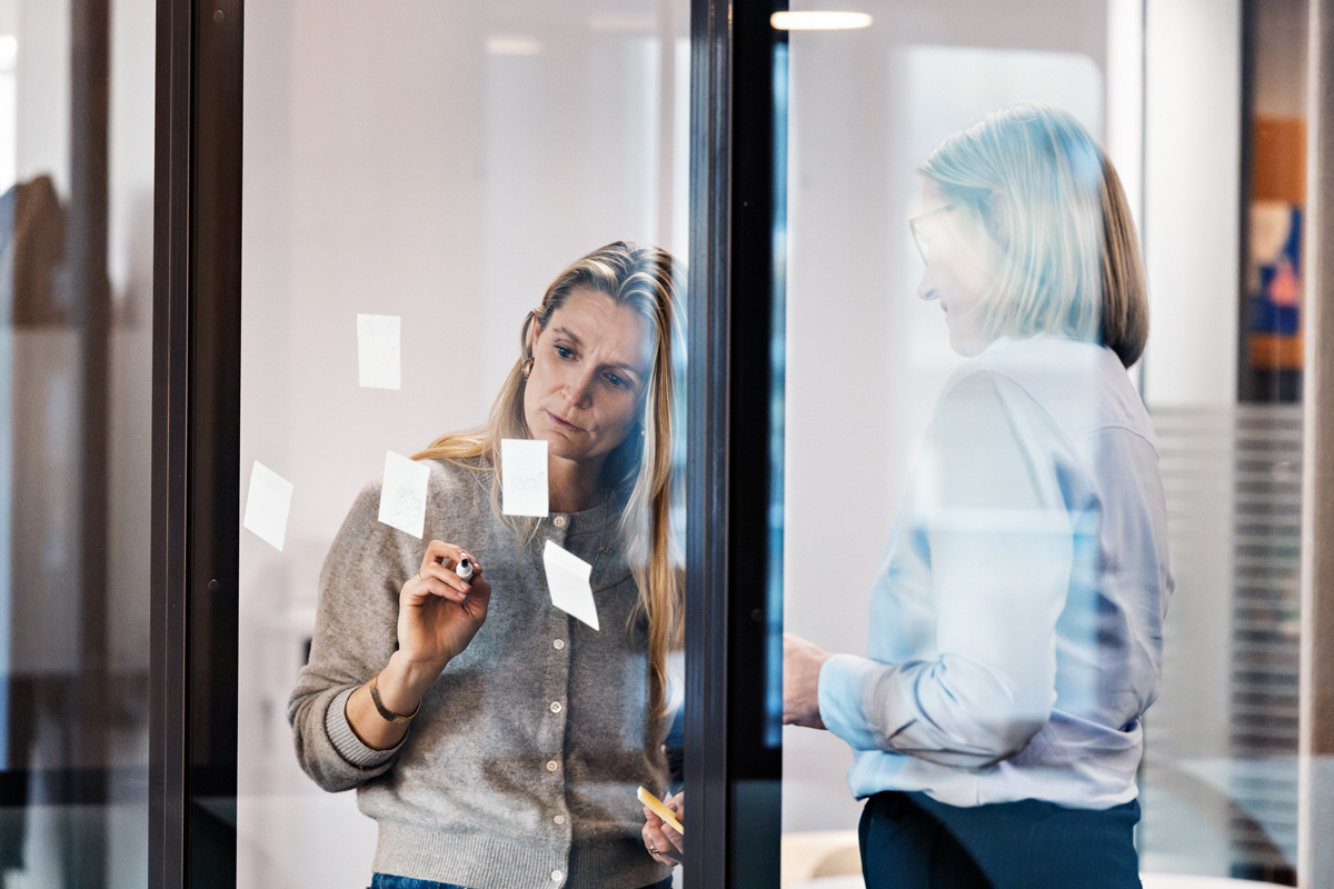 To kvinner jobber inne på et grupperom med glassvegger der de henger opp post-it-lapper