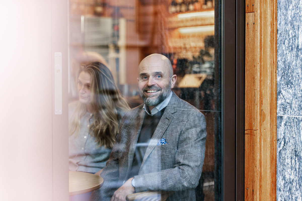En mann i grå dress sitter rundt et bord med flere inne på en kafé og ser ut av vinduet