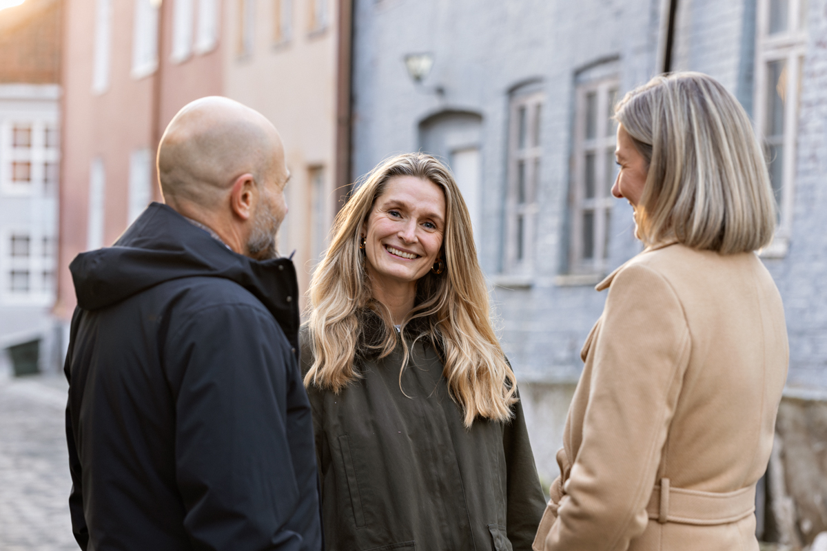 Tre personer står ute på gaten og prater, den ene smiler til kamera