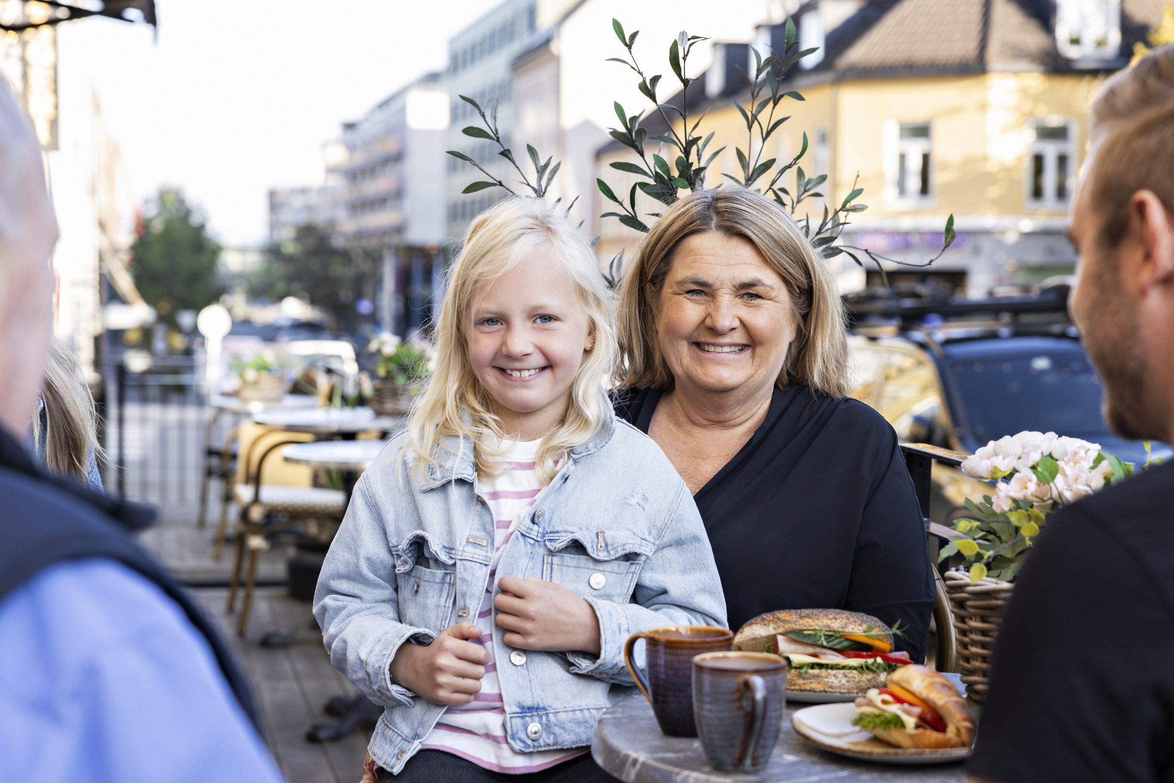 En ung jente og en kvinne sitter ute på en kafé på en solfylt dag, begge smiler til kamera