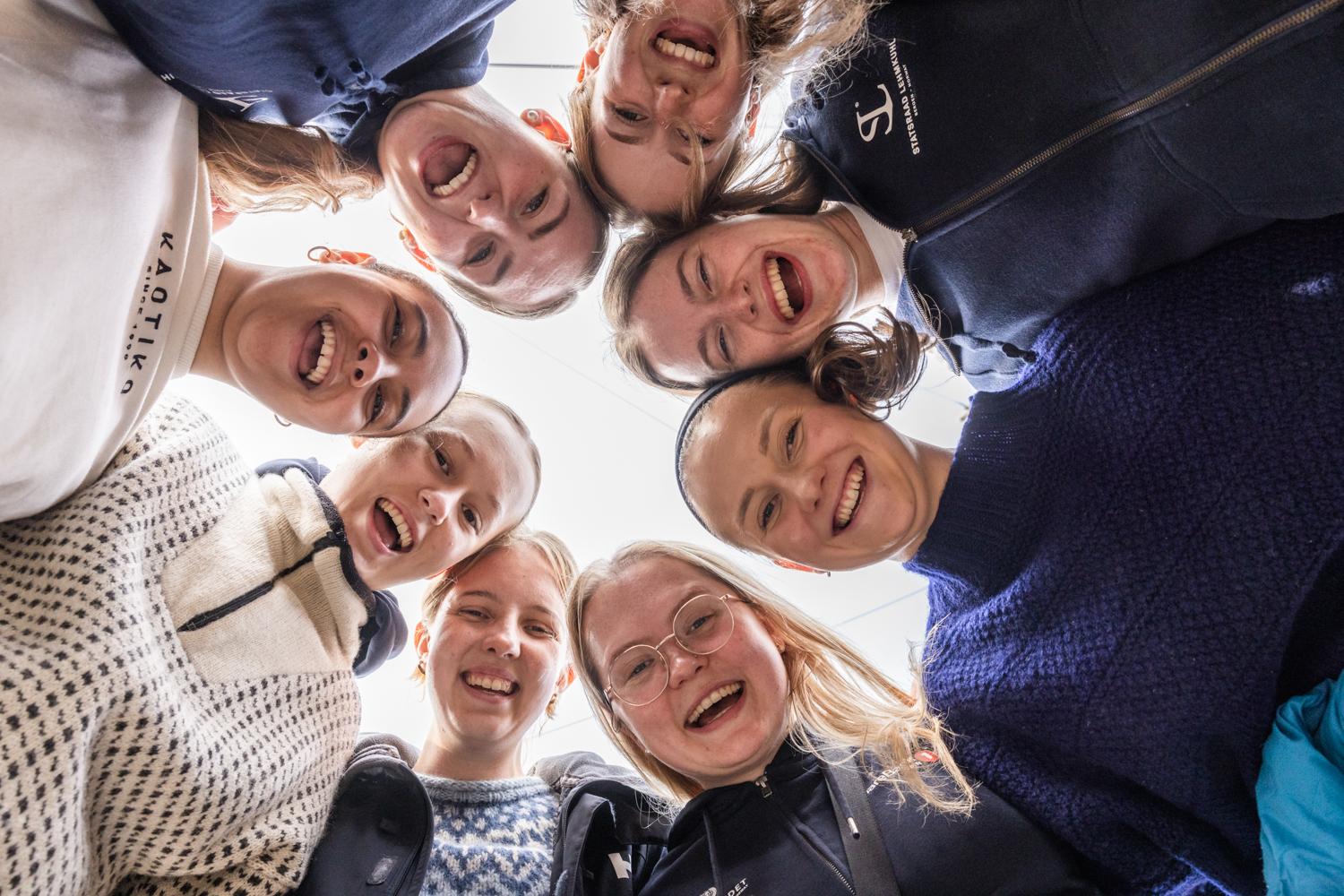 Åtte smilende unge kvinner står i en sirkel og ser ned i kamera i midten