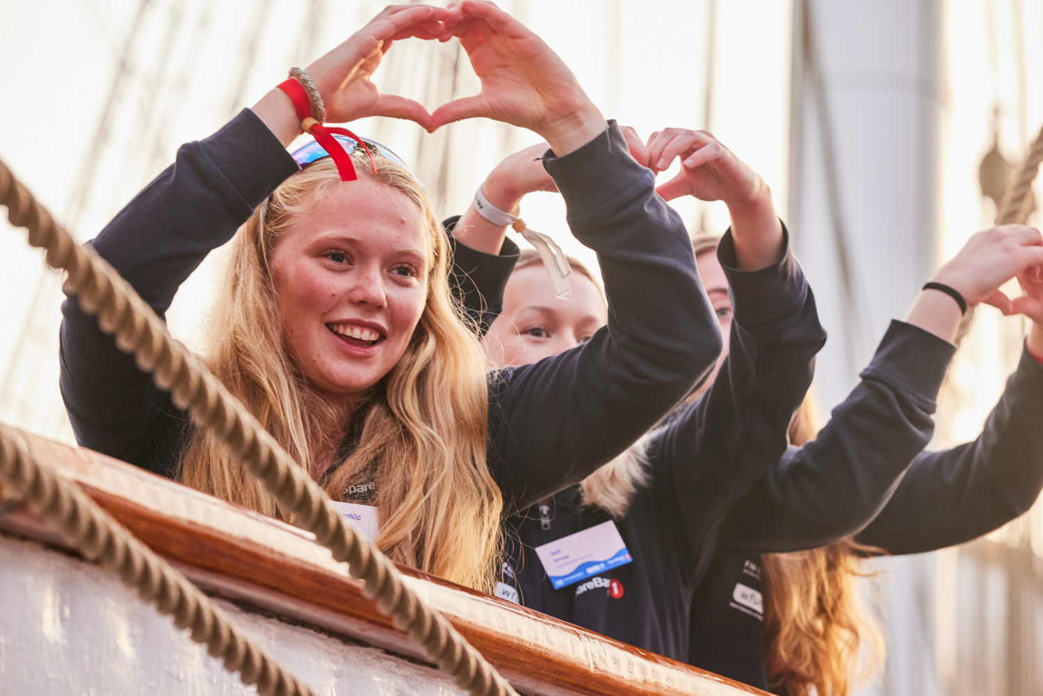 Tre unge kvinner former hvert sitt hjerte med fingrene som de holder opp over hodet mens de ser over en båtkant