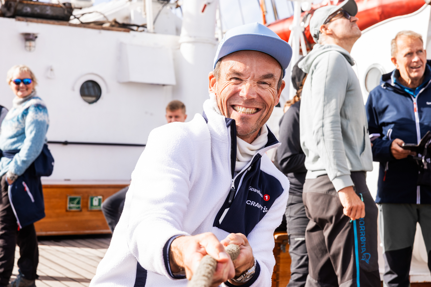 En mann iført lyse klær står og trekker tau på en kai mens han smiler til kamera