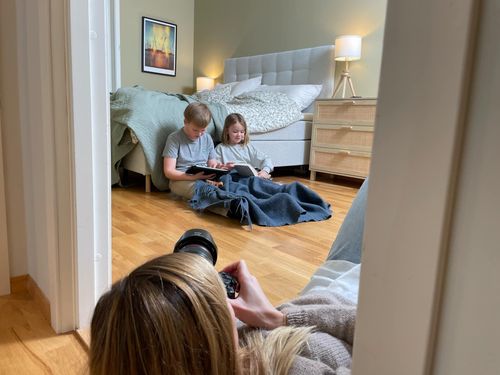 To barn leser bok og ser på ipad mens de sitter på gulvet lent mot en seng, i forgrunnen ser vi en kvinne som tar bilde av dem