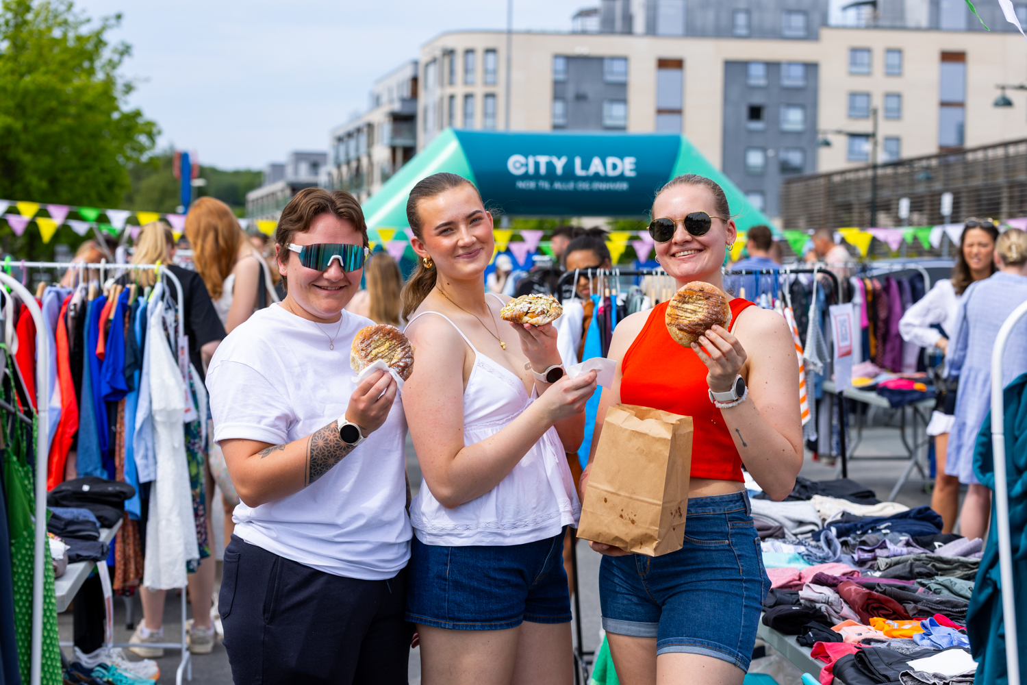 Tre unge kvinner står ute med hver sin bolle, bak dem et sort bruktmarked