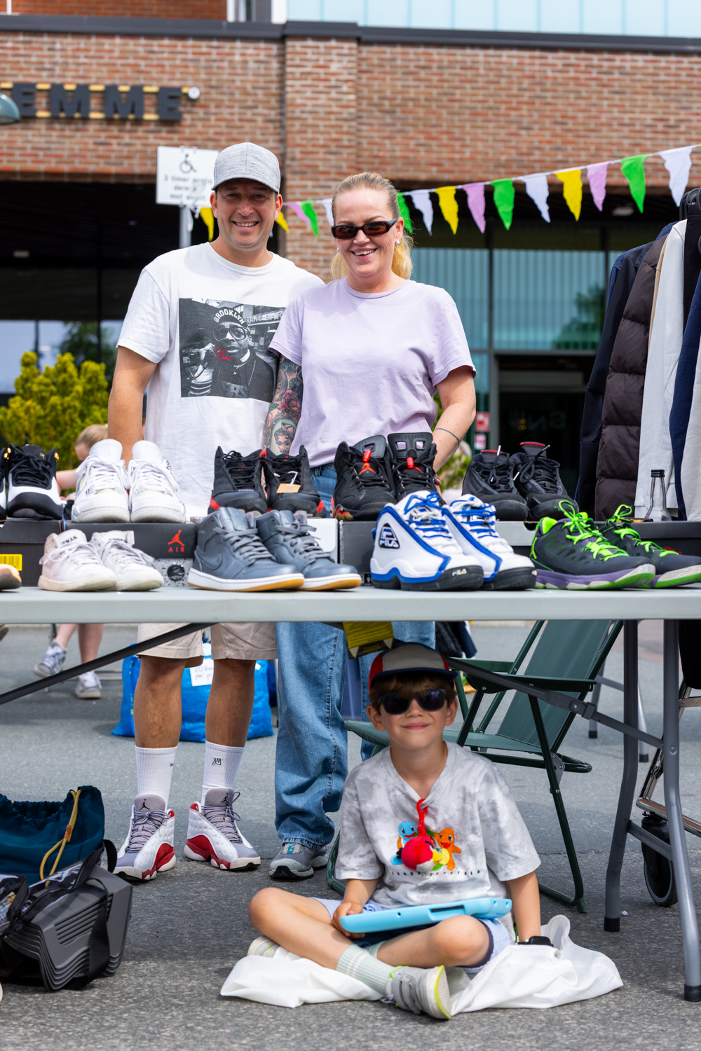 En mann og kvinne står bak en plastbord på et bruktmarked og selger ulike brukte sko, en liten gutt sitter under bordet og smiler iført caps og solbriller