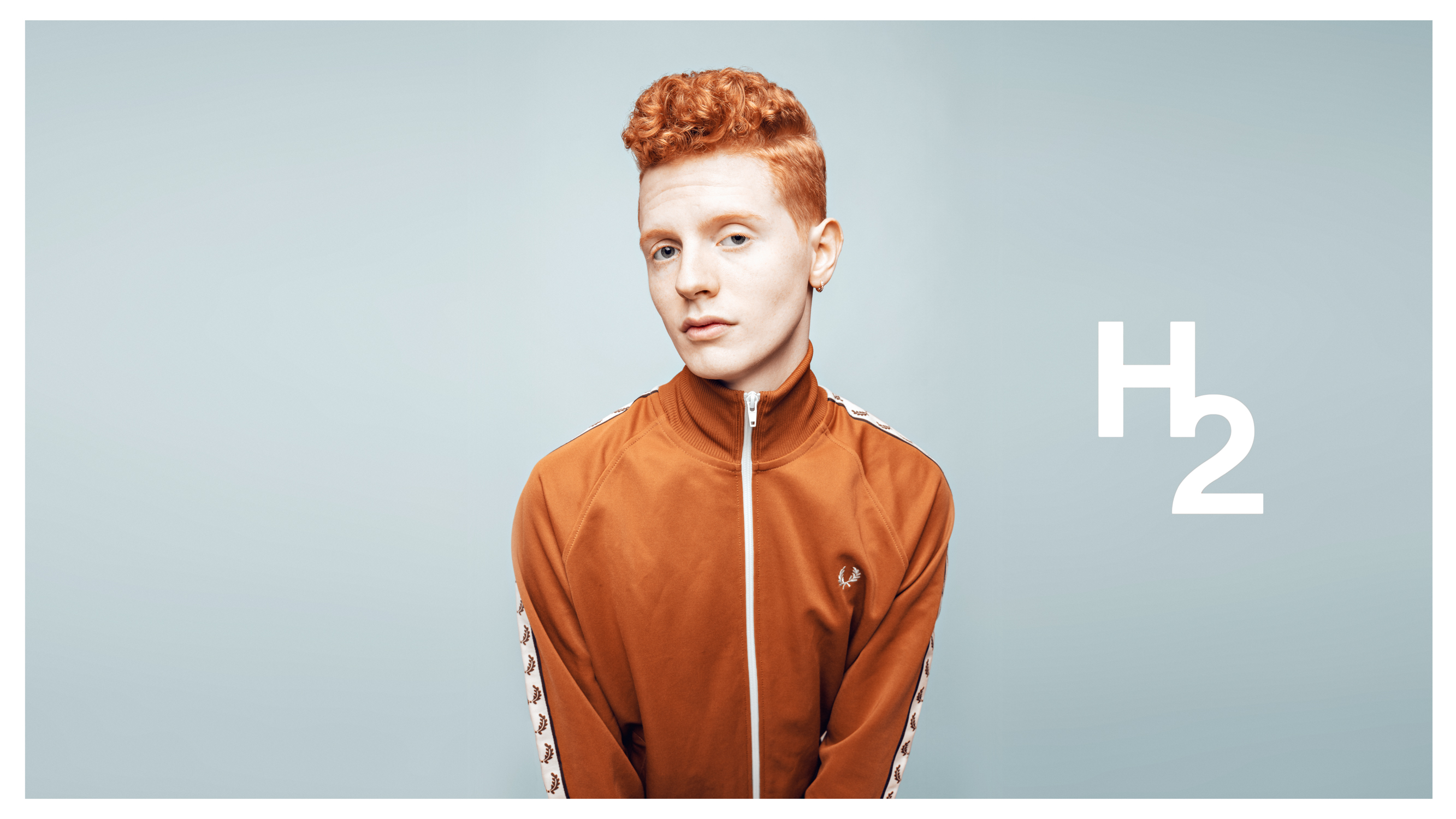 En ung mann med rødt hår poserer og ser i kamera i et duseblått studio