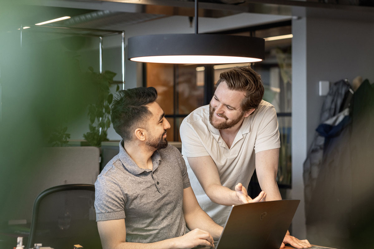 To unge menn ser på hverandre og smiler inne på et møterom
