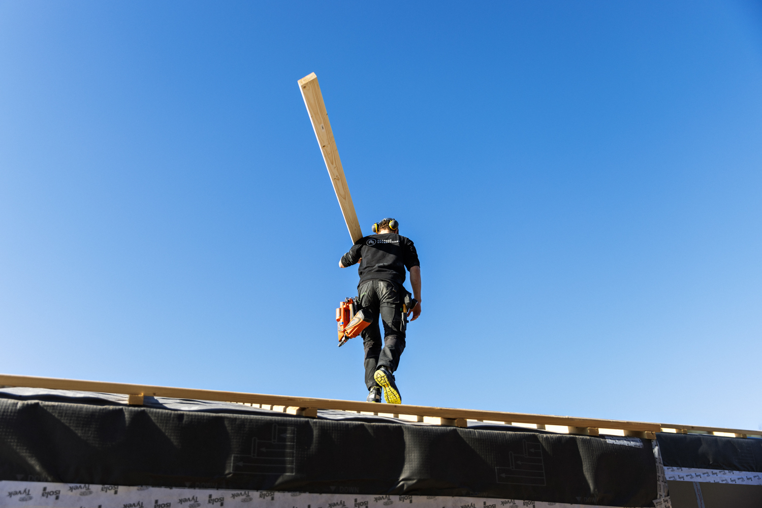 En tømrer løfter en planke på skulderen mens han går oppå et tak
