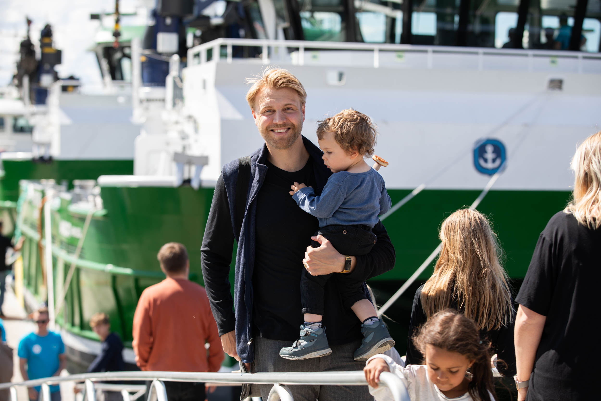 En mann holder et barn i hendene på en havn, med en stor hvit og grønn båt i bakgrunnen