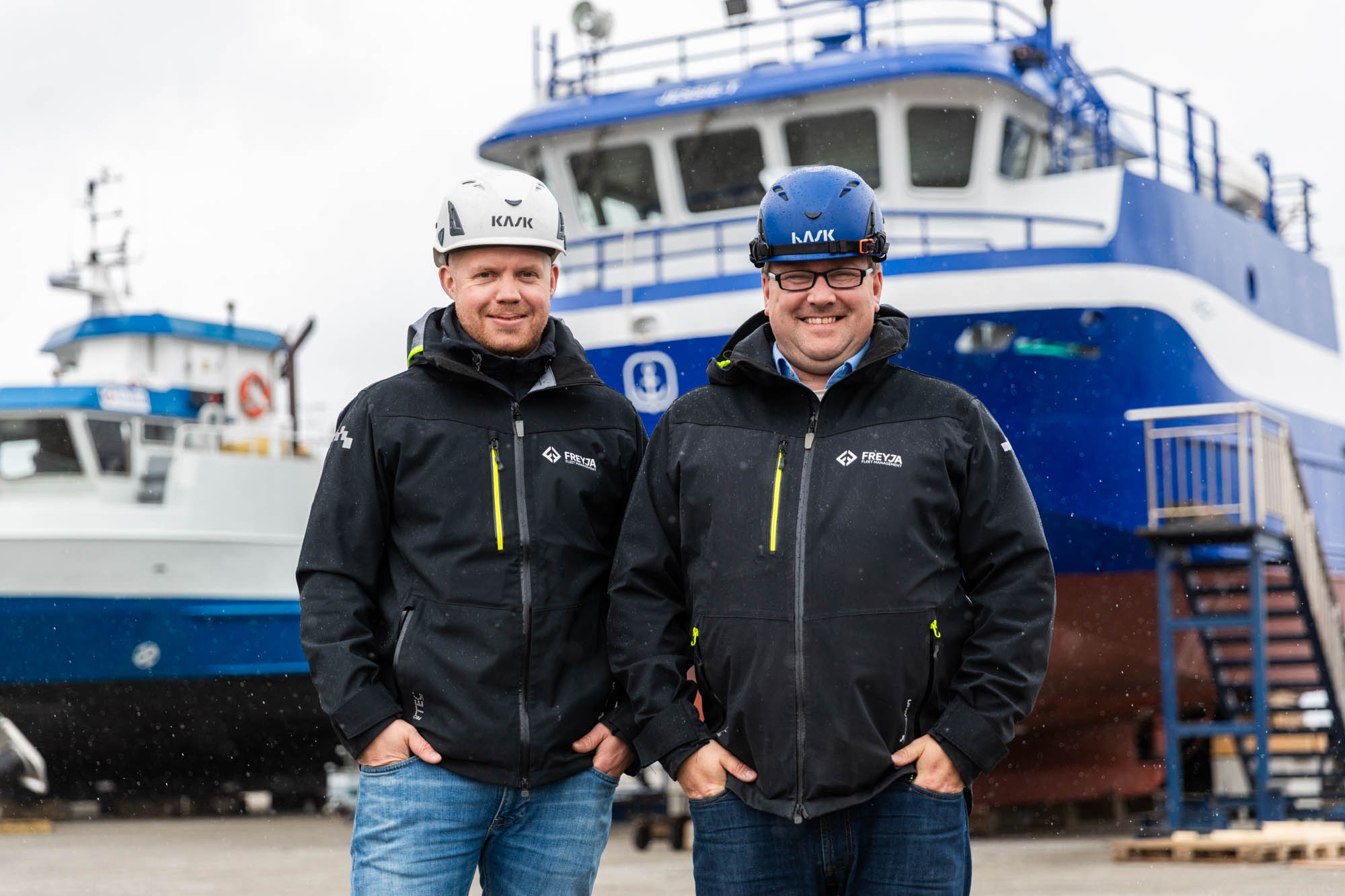 To smilende menn som står på en havn foran en båt