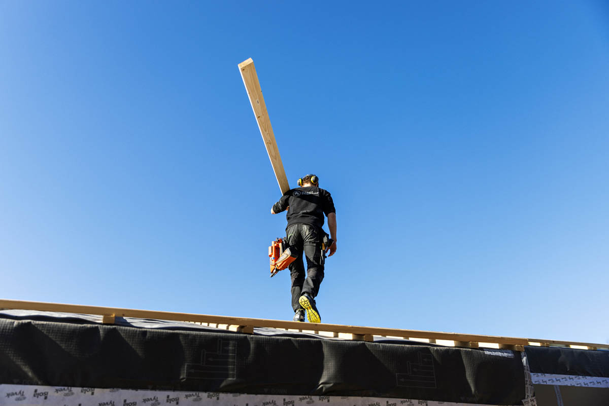 En tømrer som løfter en lang treplanke, blå himmel i bakgrunn, utsnitt tatt i froskeperspektiv.